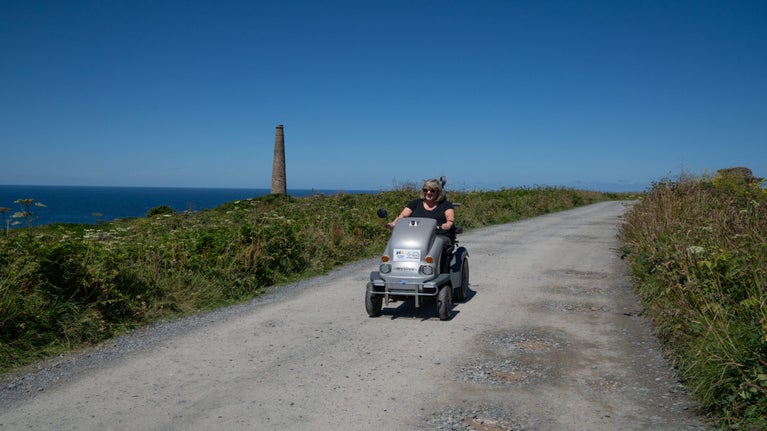 A person driving a mobility scooter along a path, with the sea and a chimney stack in the background, Botallack ,Cornwall.